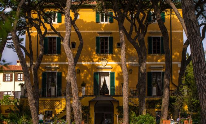 Villa gialla con persiane verdi, alberi e giardino fiorito.